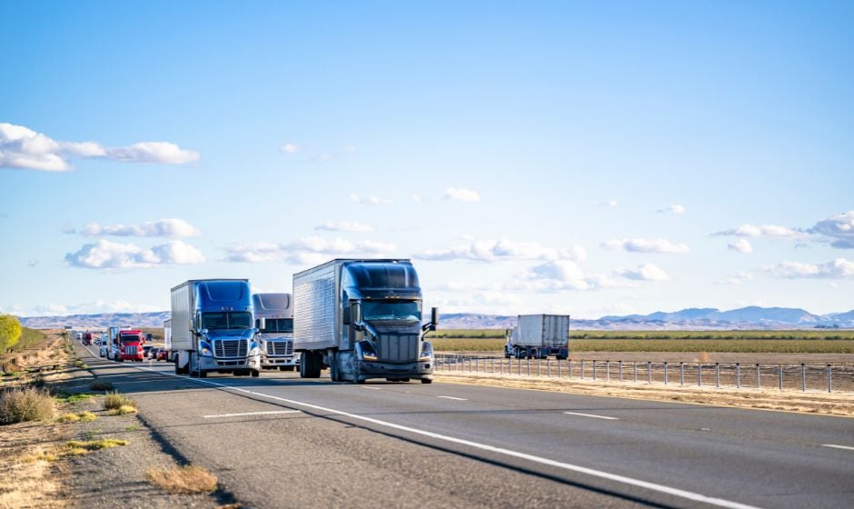 Several semi trucks with van trailers travel down a remote road