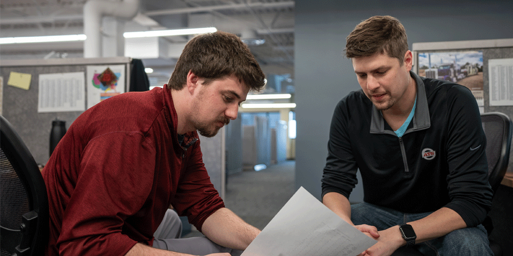 Two men on office chairs talking closely over a document.