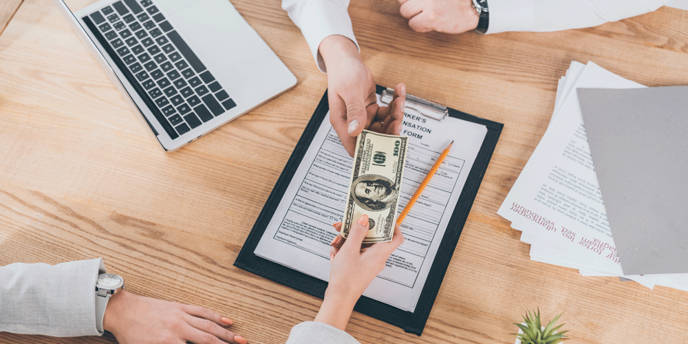 Two people pass a $100 bill at a desk. Compensation package concept.