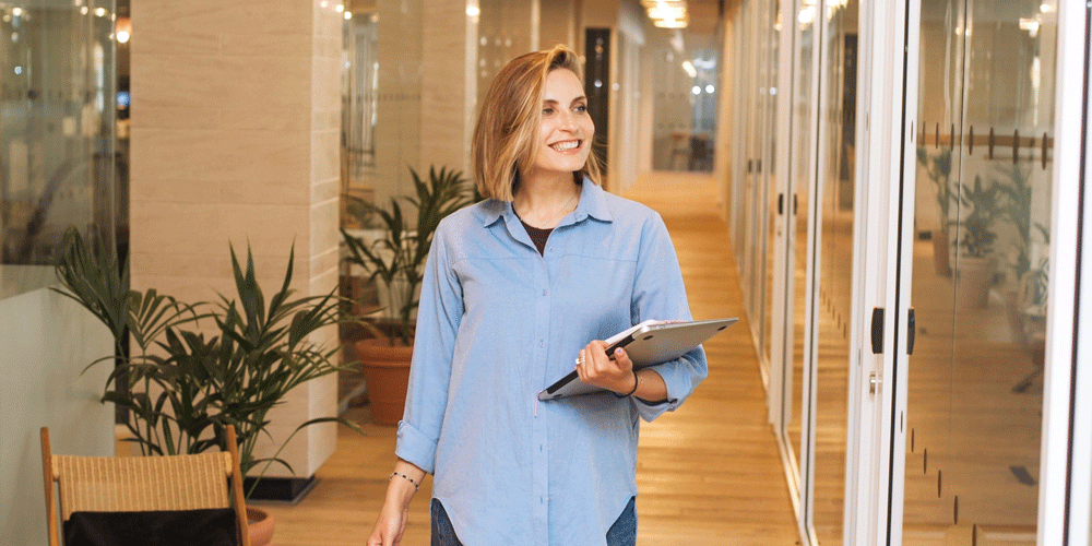 Smiling woman walking through the workplace hallway holding a laptop and notebook.