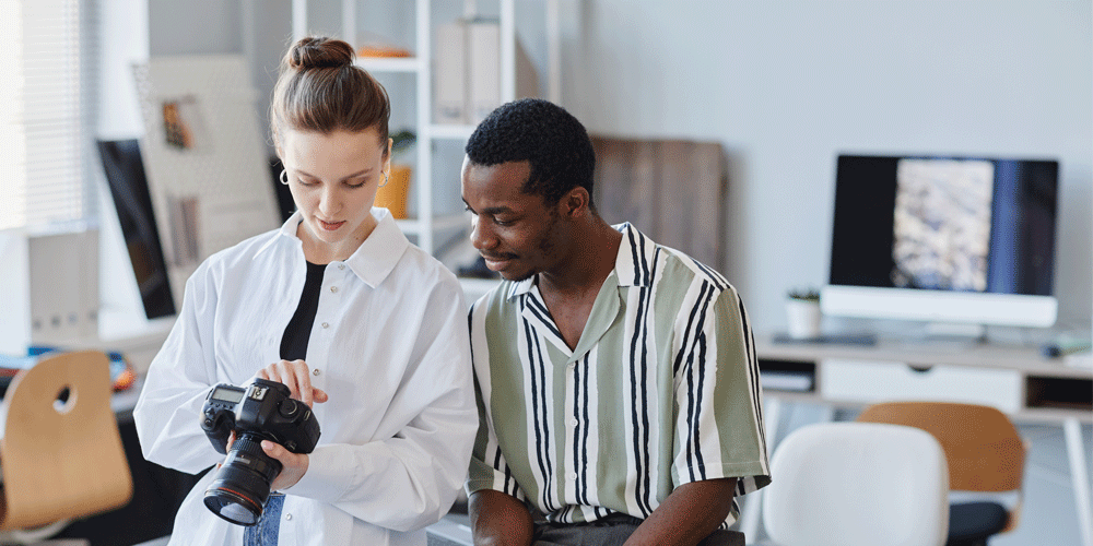 Two young, trendy interns perched on the edge of a desk reviewing footage on a camera.